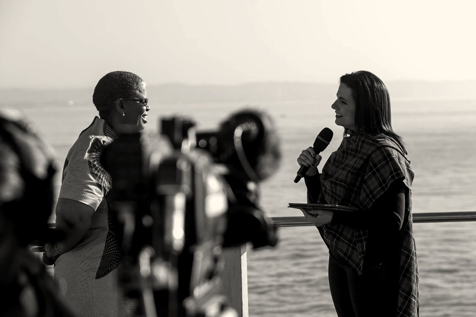 A broadcast camera in the foreground filming a presenter interviewing a guest beside the sea