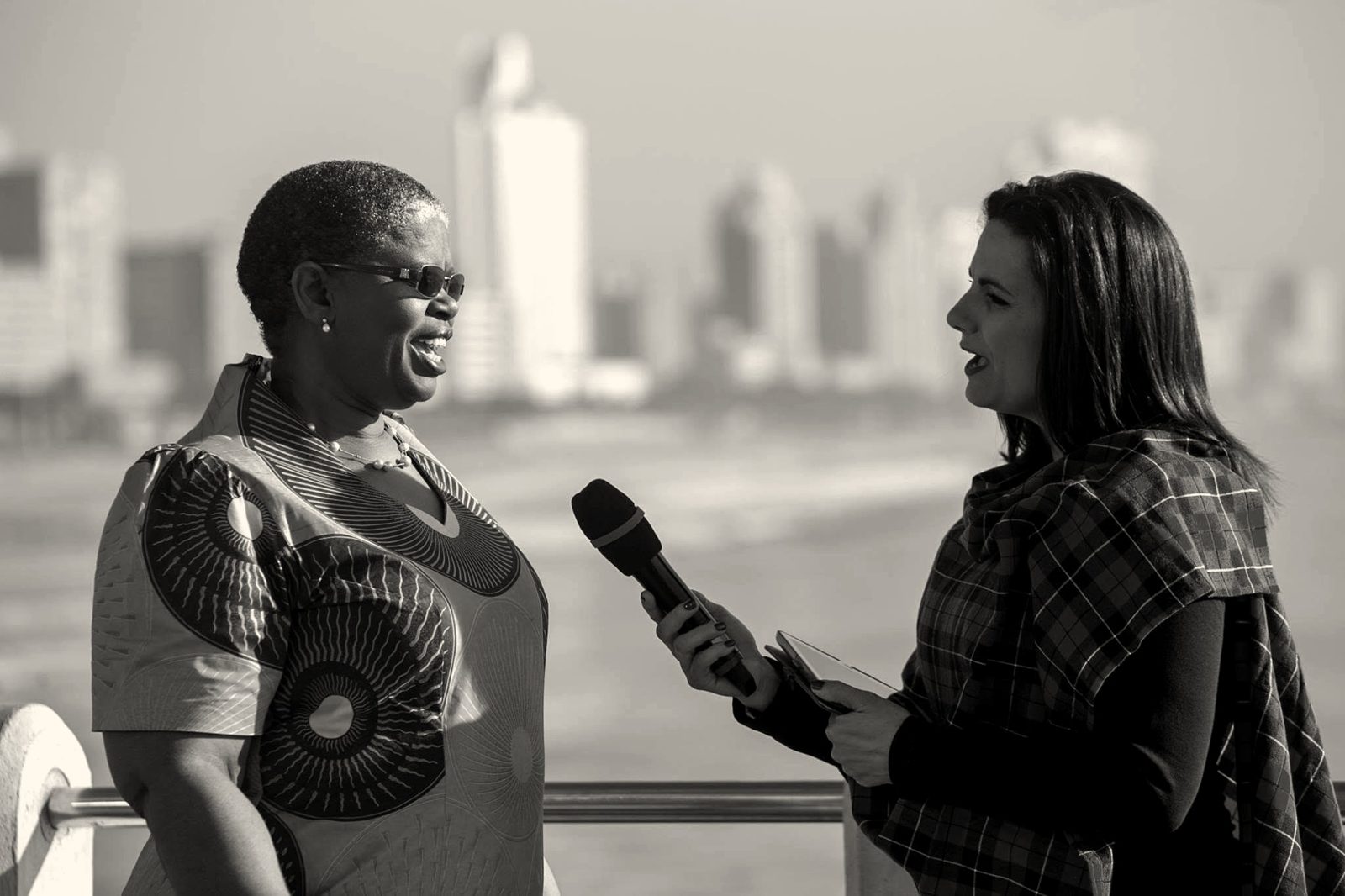 A presenter with a microphone interviewing a guest on a promenade with a city skyline behind
