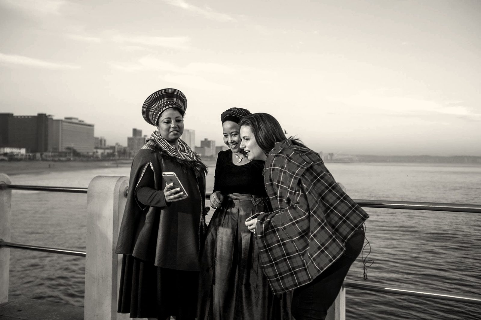Three women looking at a phone on a pier at dusk