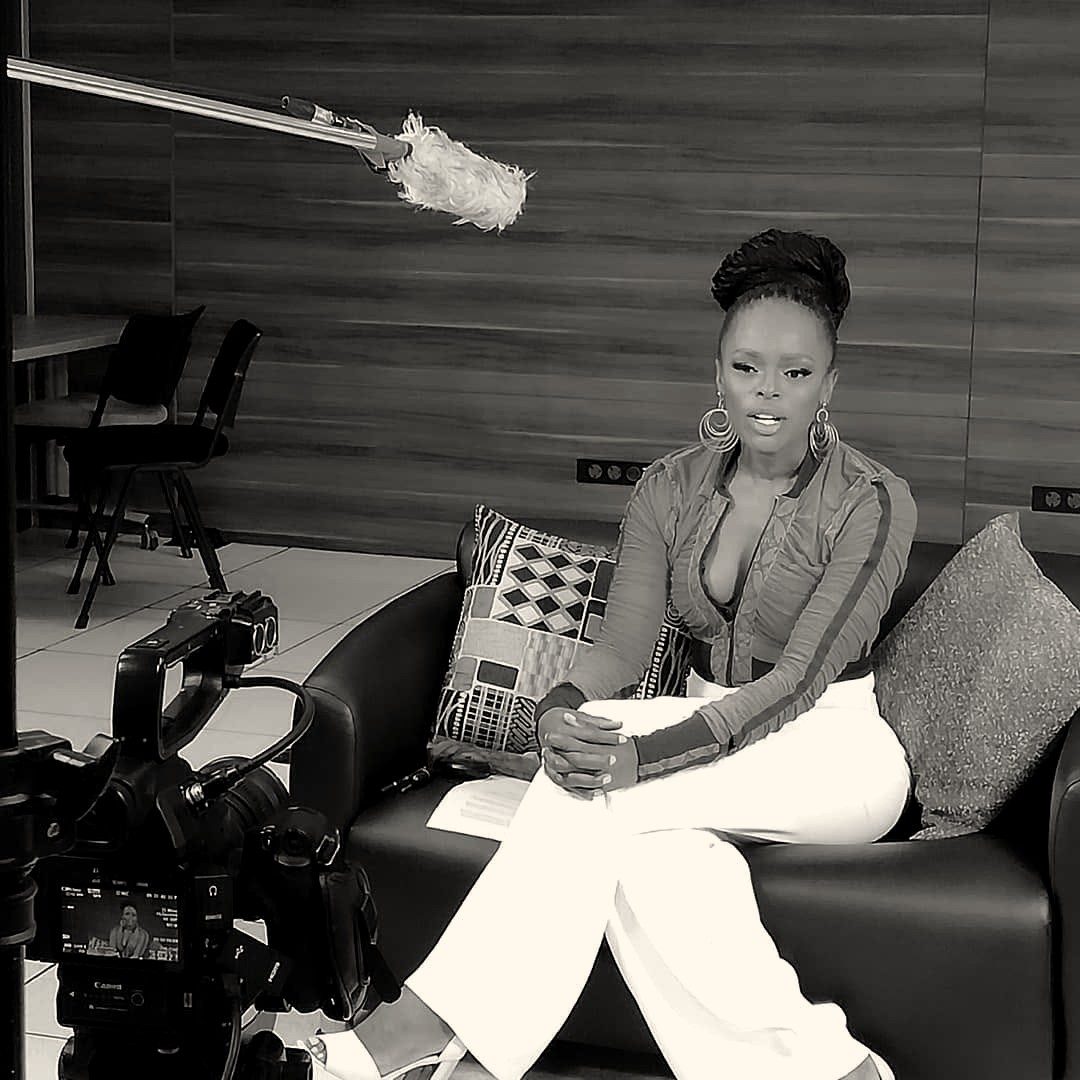 A presenter seated on a sofa being filmed, boom microphone overhead