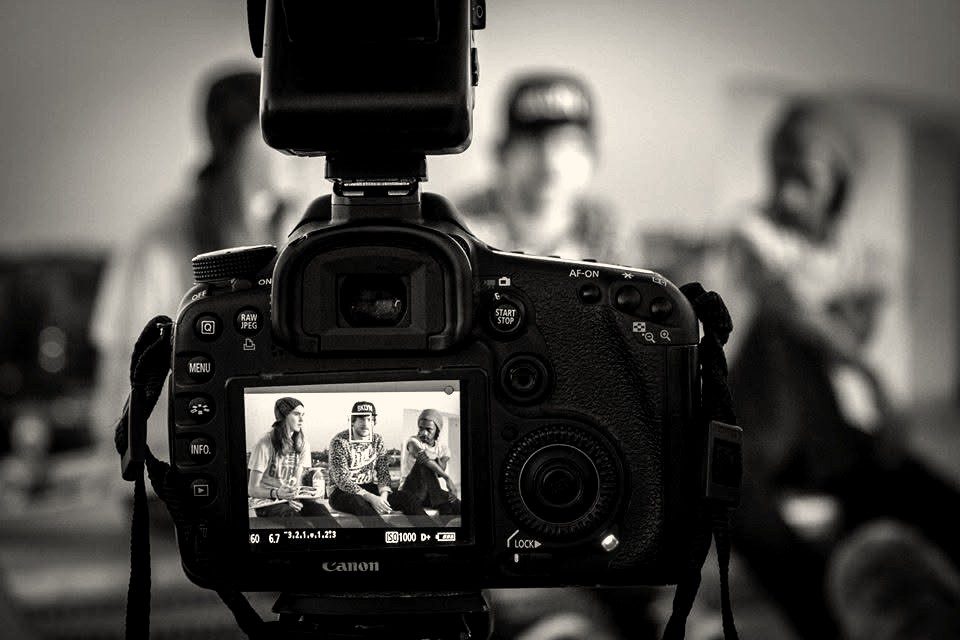 A camera on set, its rear screen showing three people being filmed
