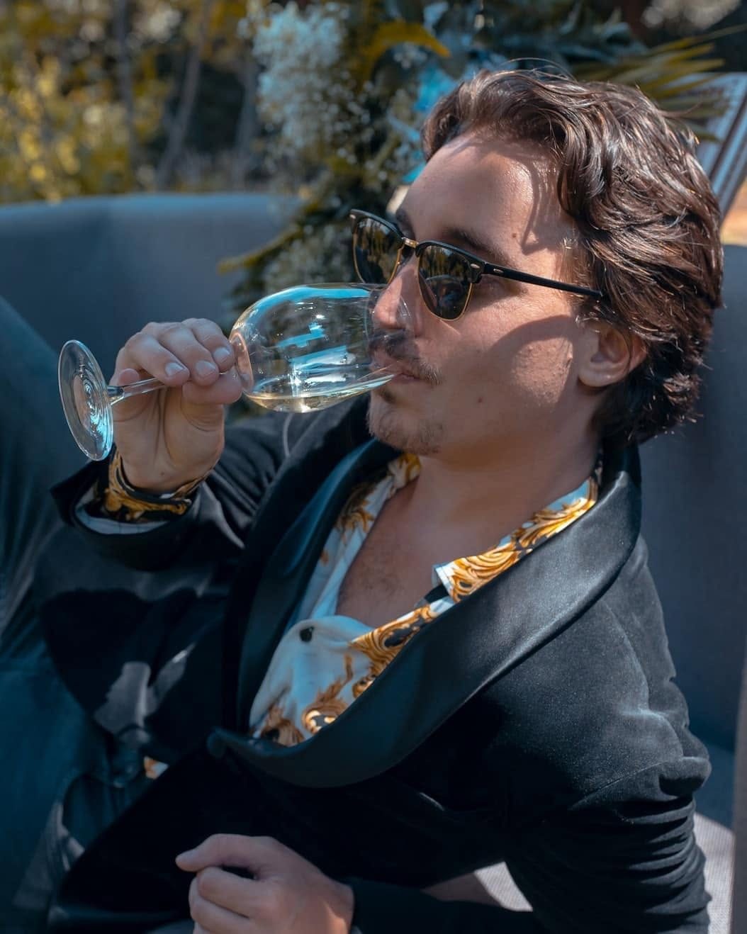 A man in sunglasses and a velvet jacket sipping white wine in a garden