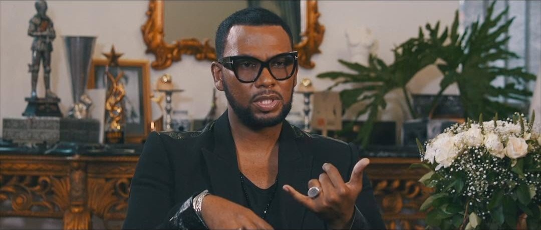 Film still: a man in black-framed glasses speaking to camera in an ornate room