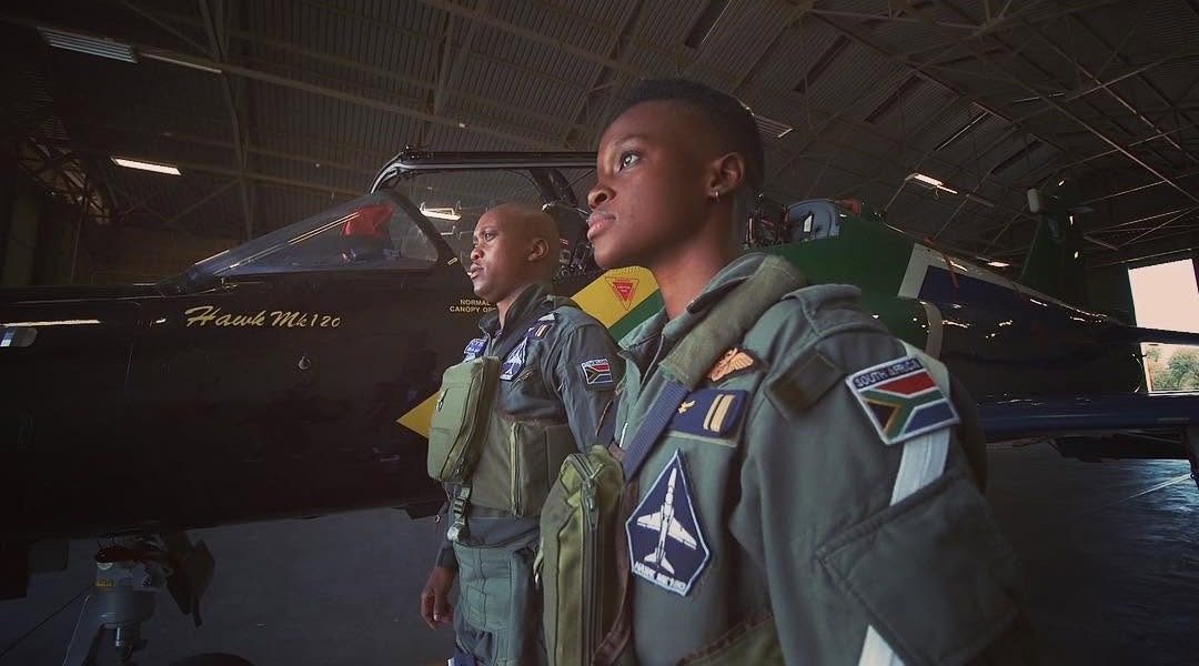 Film still: two pilots in flight suits standing beside a jet in a hangar
