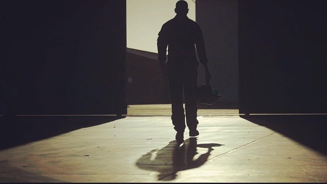 Film still: a pilot in silhouette walking out of a hangar carrying a helmet